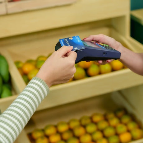 A customer holding a credit card while tapping a payment terminal. The customer is standing in a grocery store beside wooden display bins containing produce.
