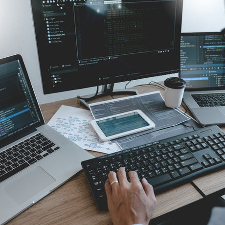 A person working at a desk with a monitor, two laptops, and a smartphone all displaying computer code.
