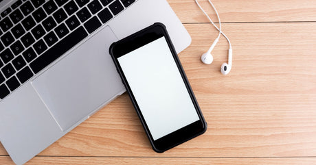 A black iPhone with a blank, white screen sits on the edge of an open MacBook. Wired headphones are next to the laptop.