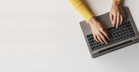An aerial view shows a person wearing a yellow sweater typing on an open laptop that sits on a white surface.