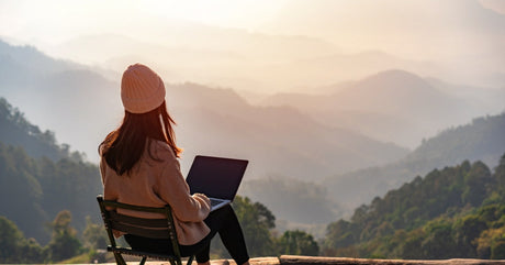 A woman sits in a small folding chair with a laptop on her lap. She sits on a cliff overlooking a mountainous landscape.