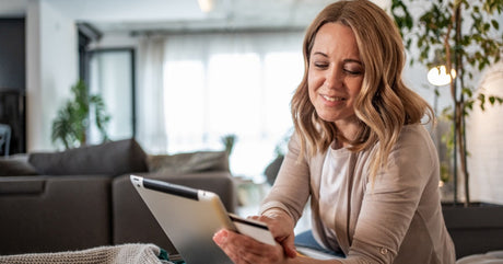 A woman is leaning on a soft blanket, holding a tablet and a credit card in her hand and scrolling with the other hand.