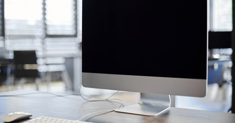 A desktop monitor sits on a wooden desk with a white keyboard and a white computer mouse. A white cord exits the keyboard.