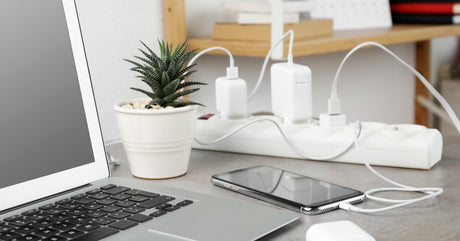 A white surge protector sits on a desk with chargers plugged in. A laptop, a cellphone, and an AirPods case sit on the desk.