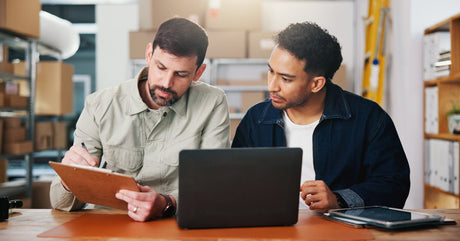 Two men are sitting at a table, looking at a clipboard that one of them is writing on. A laptop is in front of them.