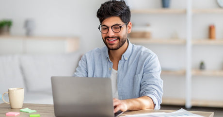 A smiling man sits at a wooden table typing on the keyboard of a laptop. Papers, a mug, and sticky notes are on the table.