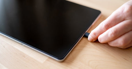 A close-up view shows a person plugging a black charging cord into the charging port of a tablet.