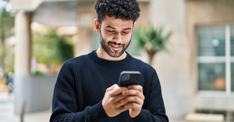 A young man wearing a dark-colored sweater is smiling and holding a smartphone in his hands. He's looking at the screen.
