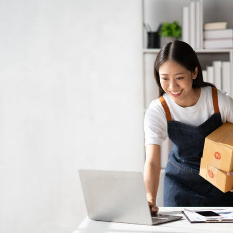 A smiling woman wearing a dark blue apron is leaning over to type on a laptop. She has a box tucked under her arm.