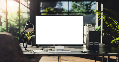A desktop monitor with a white screen sits on a desk next to a lamp, a keyboard, a notebook, and a computer mouse.