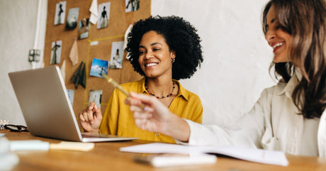Two businesswomen sit at a wooden table looking at a laptop and smiling. A corkboard is on the wall behind them.
