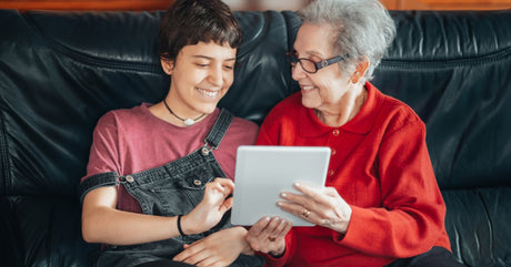A senior adult woman sits on a black leather couch next to a young woman looking at an iPad. They are both smiling.