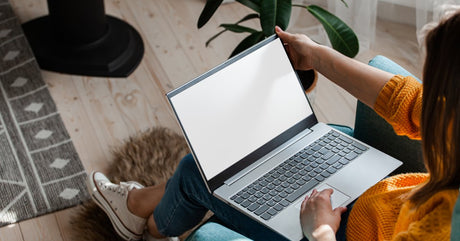 A woman wearing an orange sweater, jeans, and white sneakers sits in a teal chair with a laptop on her lap.