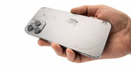 A close-up view shows a person's hand holding a cracked silver iPhone in their hand with the screen facing their palm.