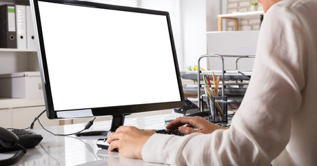 A person wearing a white cardigan sits at a white desk typing on a black keyboard of a desktop PC.