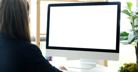 A woman wearing a dark-colored shirt sits at a desk looking at a blank monitor. Plants appear around the desk.