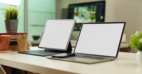 A laptop, tablet with a keyboard, and smartphone rest on a white desk with a blurred background and office supplies.