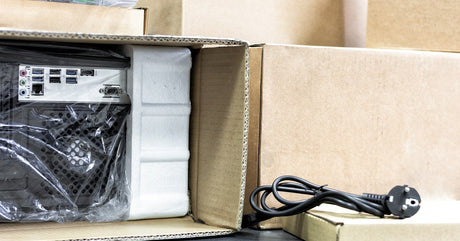 A packaged computer tower in a cardboard box with foam padding, alongside a black power cable and stacked brown boxes.