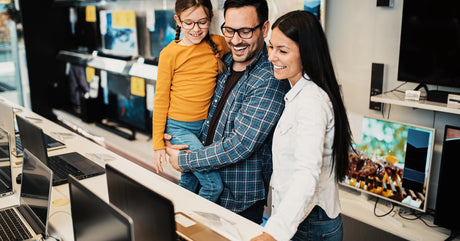 A man in glasses holds a young girl while smiling with a woman in a white shirt as they browse laptops in a store.