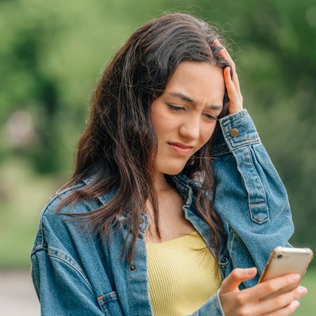 A young woman wearing a denim jacket is holding a smartphone in one hand and touching her head with the other.