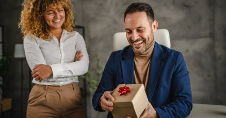 A man is smiling at a gift box with a red bow while a woman stands nearby with her arms crossed in an office.
