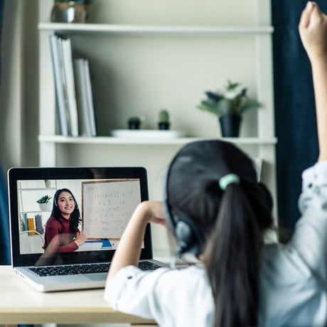 A girl in headphones is raising her hand at a desk while attending an online class on a laptop showing a female teacher.