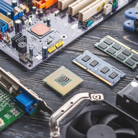 Computer parts showing RAM, CPU, fans, and drives resting on a dark surface beside a scattered layout of circuit boards.