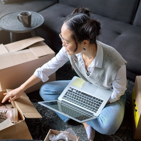 A woman holding a laptop while reaching into a cardboard box with tan boots beside her on the carpet.