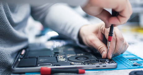 A close-up of a technician using a precision screwdriver to repair a disassembled laptop on a blue mat.