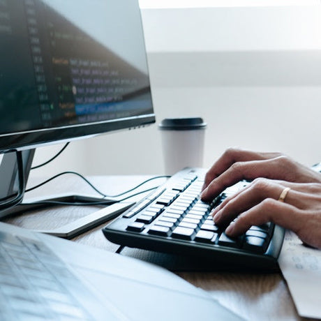 A person in a long-sleeve blue shirt typing on a black keyboard between two monitors and a laptop on the desk.