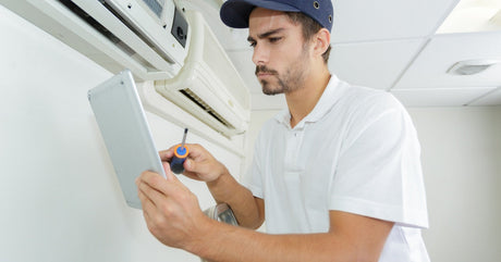A man in a white polo shirt using a screwdriver and iPad while standing near a wall-mounted air conditioner.