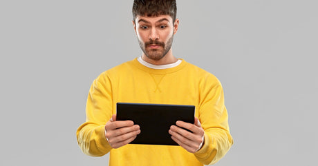 A man in a yellow sweatshirt looking at a tablet with a surprised expression against a plain gray background.