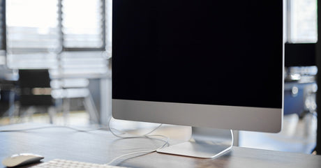 Modern desktop computer with a blank black screen, keyboard, and mouse on a wooden desk in a bright office setting.