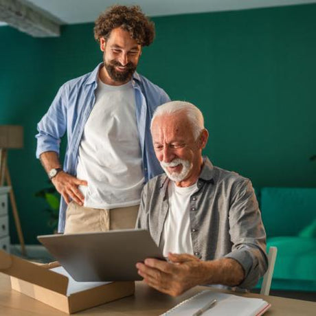 An elderly man smiling joyously and unboxing a new laptop, while a man is standing beside him inside a home.
