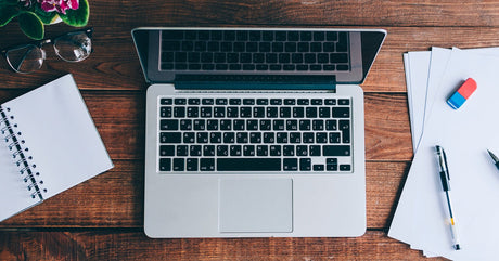 A top view of an open laptop on wooden desk with a notebook, pen, papers, eraser, and eyeglasses arranged around it.