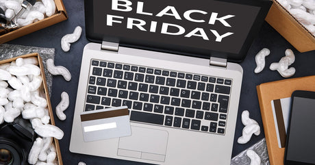 An open laptop showing "Black Friday" text on-screen. A credit card, packing peanuts, and shopping items lay around it.