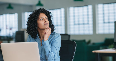 A woman sitting at a desk with a laptop as she stares off into the distance. Her hand is touching her chin, making a thinking gesture.