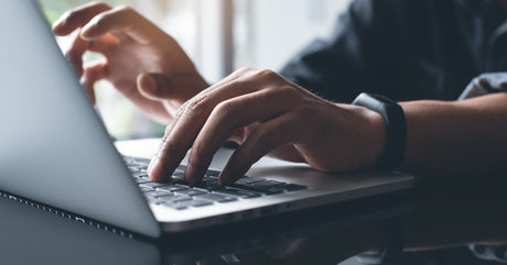 A close-up view shows a person sitting at a black desk typing on an open laptop. The person is wearing a black watch.