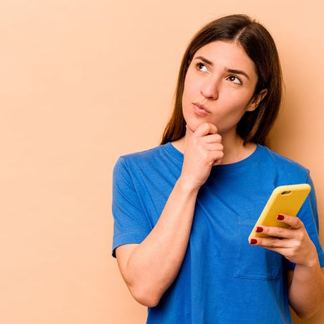 A young woman holds an iPhone in one hand while placing the other hand on her chin with a contemplative look on her face.