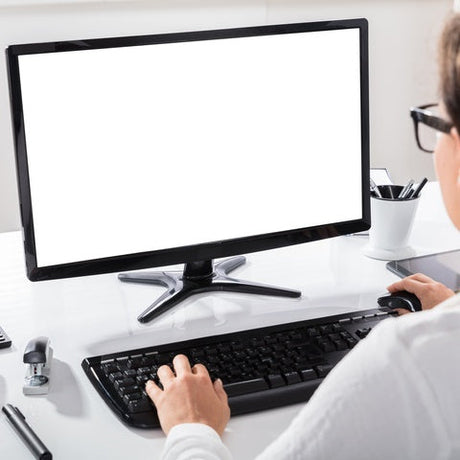 A woman sits at a white desk using a black mouse and typing on a black keyboard of a desktop computer.