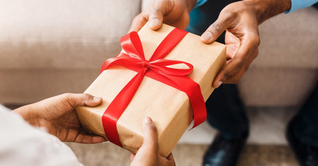 A close-up view shows two people holding a wrapped gift. The gift is wrapped in brown paper and has a red ribbon bow.