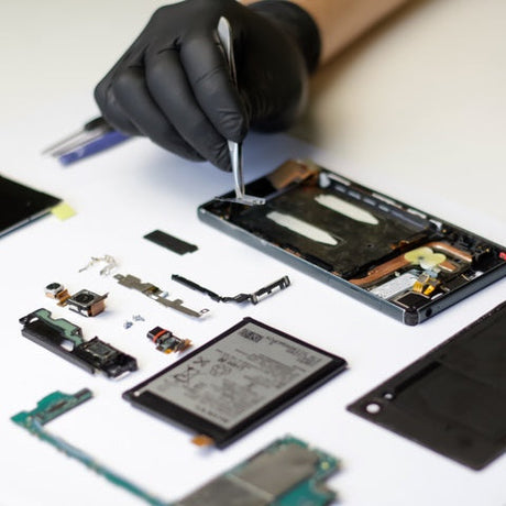 A person wearing a black latex glove is holding a pair of tweezers to work on a cellphone. The cellphone is in pieces.