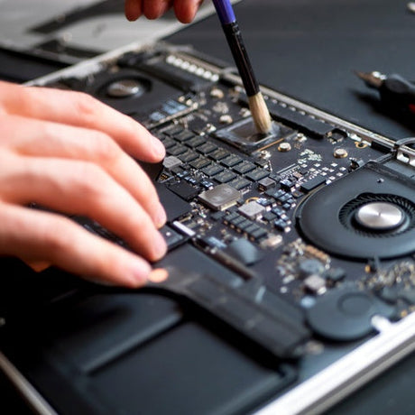 A close-up view shows a person using a small black and blue paintbrush to clean the inside of a laptop.