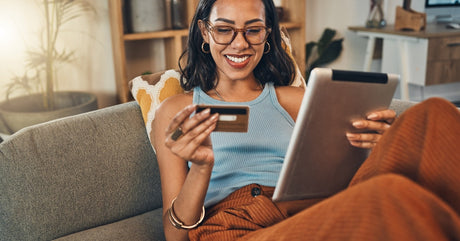 A smiling young woman lies on a couch holding a blue credit card in one hand and a silver tablet in the other.