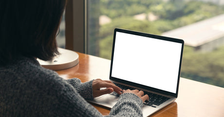 A woman wearing a speckled gray sweater sits at a wooden desk in front of a window, typing on an open laptop.