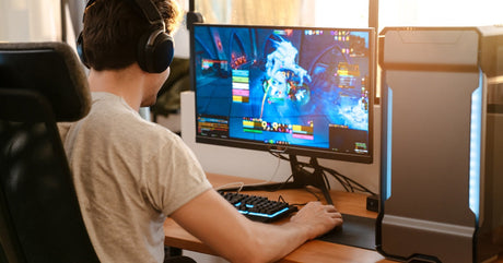 A young man sits in a black chair at a wooden desk, playing a game on a gaming computer. He's wearing black headphones.