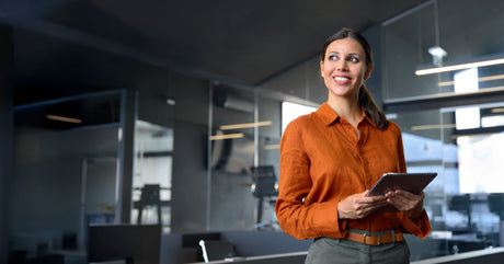 A smiling woman wearing an orange blouse and her hair in a ponytail stands in a conference room holding a tablet.