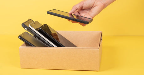 A person's hand is placing a black smartphone into a small cardboard box with other smartphones.