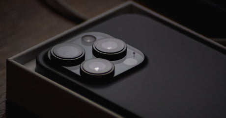 A close-up view shows a black iPhone sitting in a black box, screen side down. The background is black.