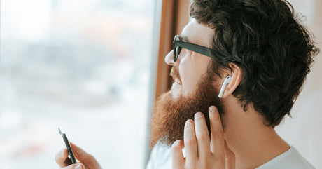 A man looks out a window, holding a black smartphone in one hand. He has a white AirPod in his ear.